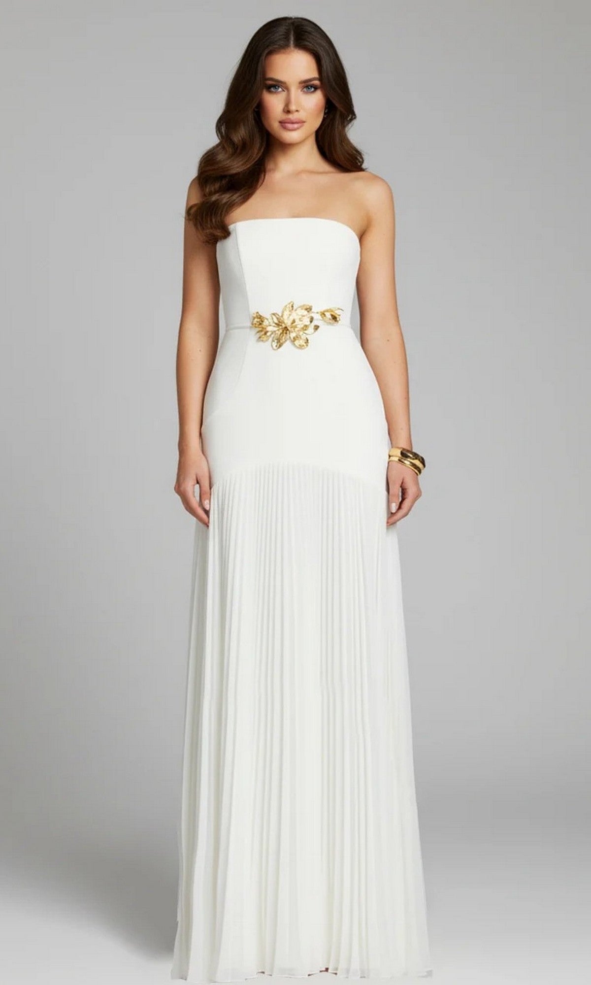 A woman stands against a plain light grey background, wearing an off-white Jovani 47000 long formal dress with a strapless pleated design, gold floral waist detail, and a gold bracelet.